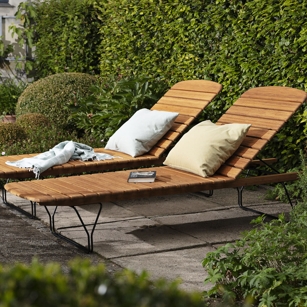 Zwei hölzerne Gartenliegen mit verstellbaren Rückenlehnen stehen auf einer Terrasse, umgeben von sorgfältig gepflegten Hecken und anderen grünen Pflanzen. Eine der Liegen hat ein helles beiges Kissen, die andere ein blasses grünes Kissen. Auf einer Liege liegt eine gefaltete Decke, auf der anderen ein Buch.