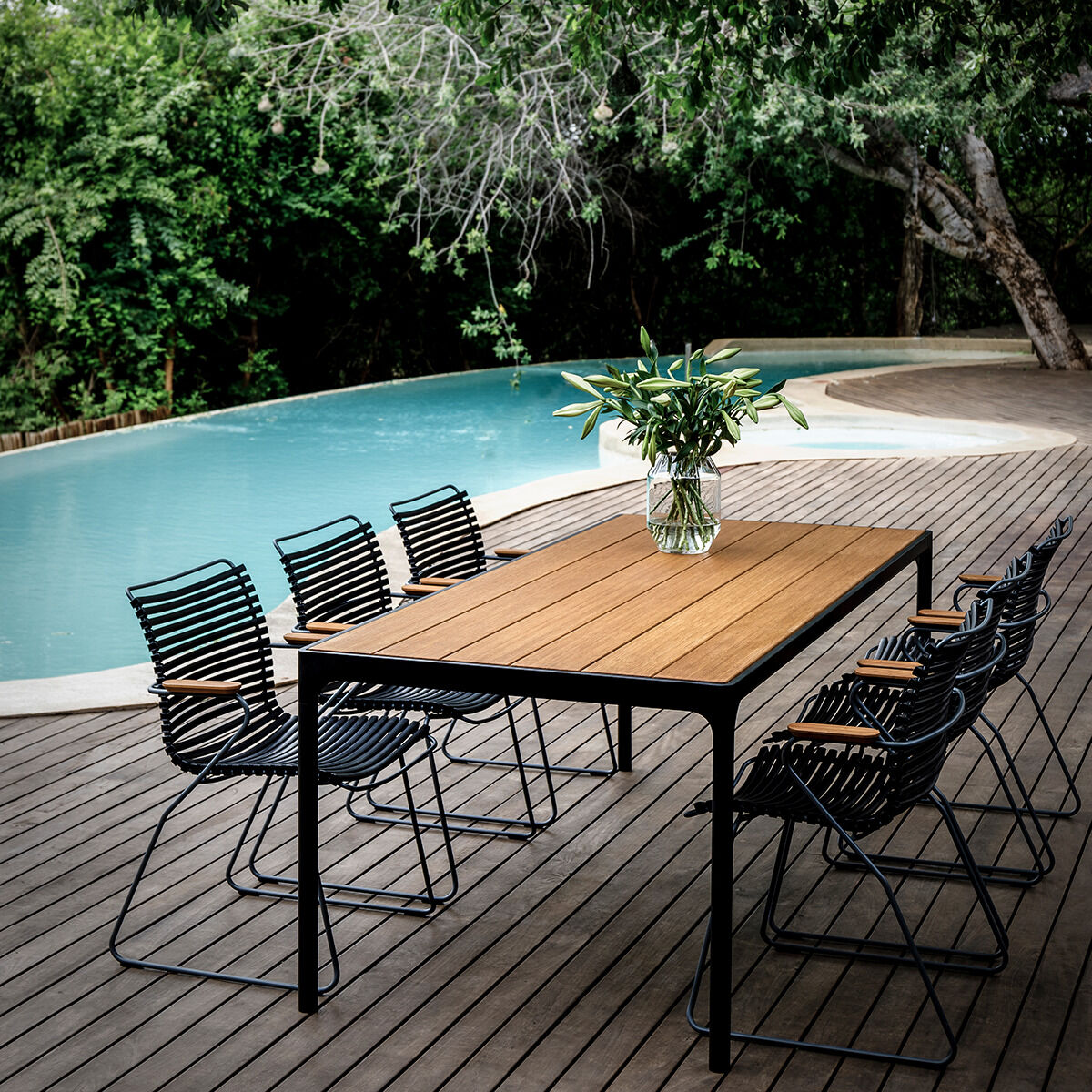 Auf einer Holzterrasse am Pool steht ein rechteckiger Esstisch mit Holzplatte und schwarzem Gestell, umgeben von sechs »CLICK Dining Chair«-Stühlen von Houe in Schwarz mit Armlehnen aus Bambus. Auf dem Tisch steht eine Vase mit frischen, grünen Blumen.