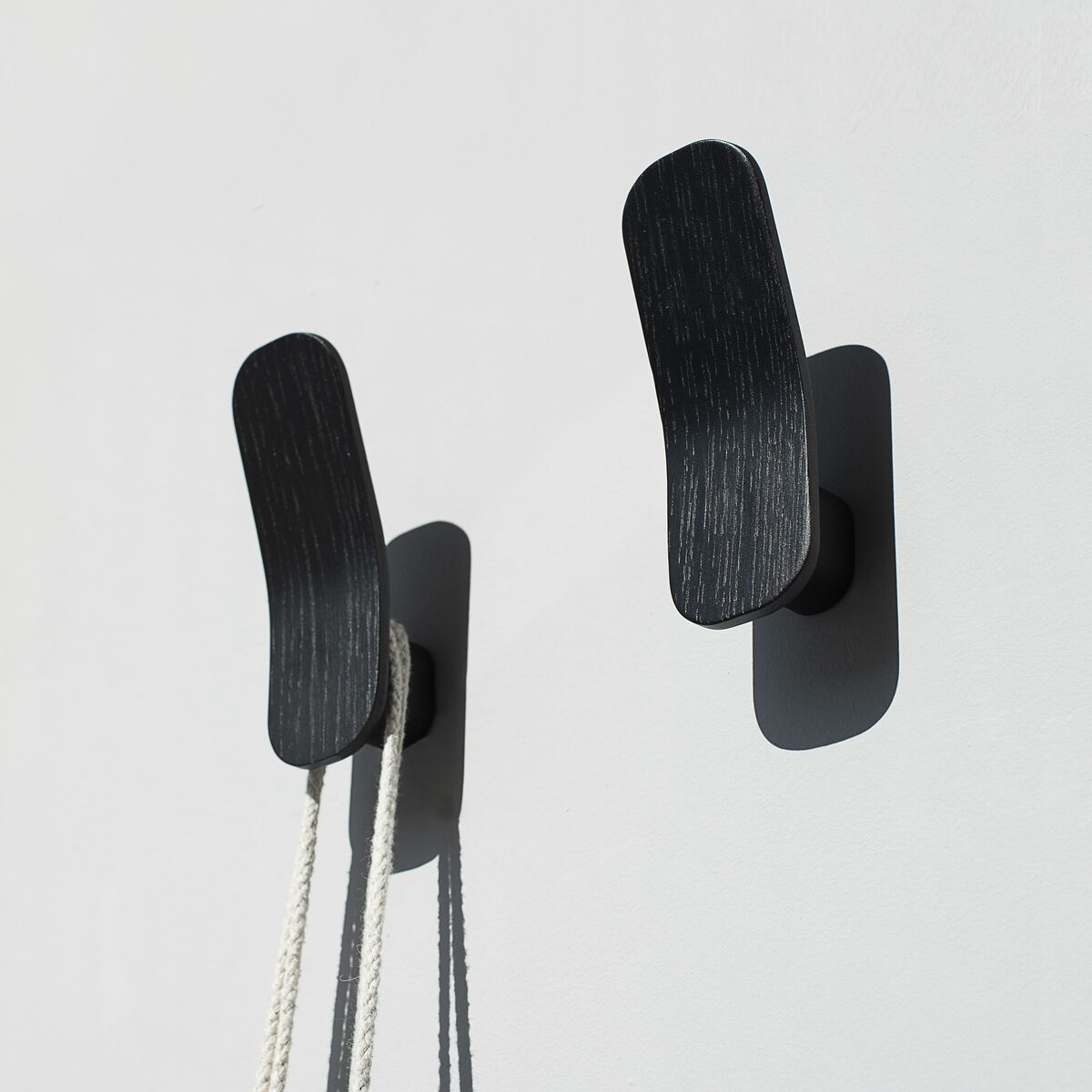Zwei moderne Kleiderhaken »Eshaku« aus schwarzer Eiche, die an einer weißen Wand montiert sind, mit einem Seil und einem schwarzen Gegenstand, der an einem der Haken hängt.