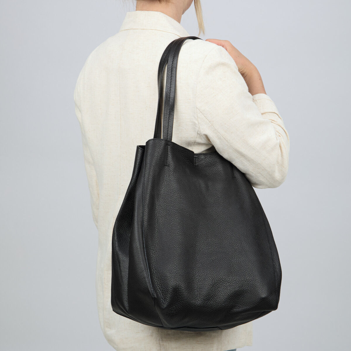 Eine Person trägt die schwarzbraune ZEIT-Tasche »Cep« aus hochwertigem Rindleder von Guerer & Guerer über der Schulter. Die Tasche hat ein reduziertes, elegantes Design, strukturierte Oberfläche und zwei lange Henkel. Sie passt perfekt zu hellen Outfits.