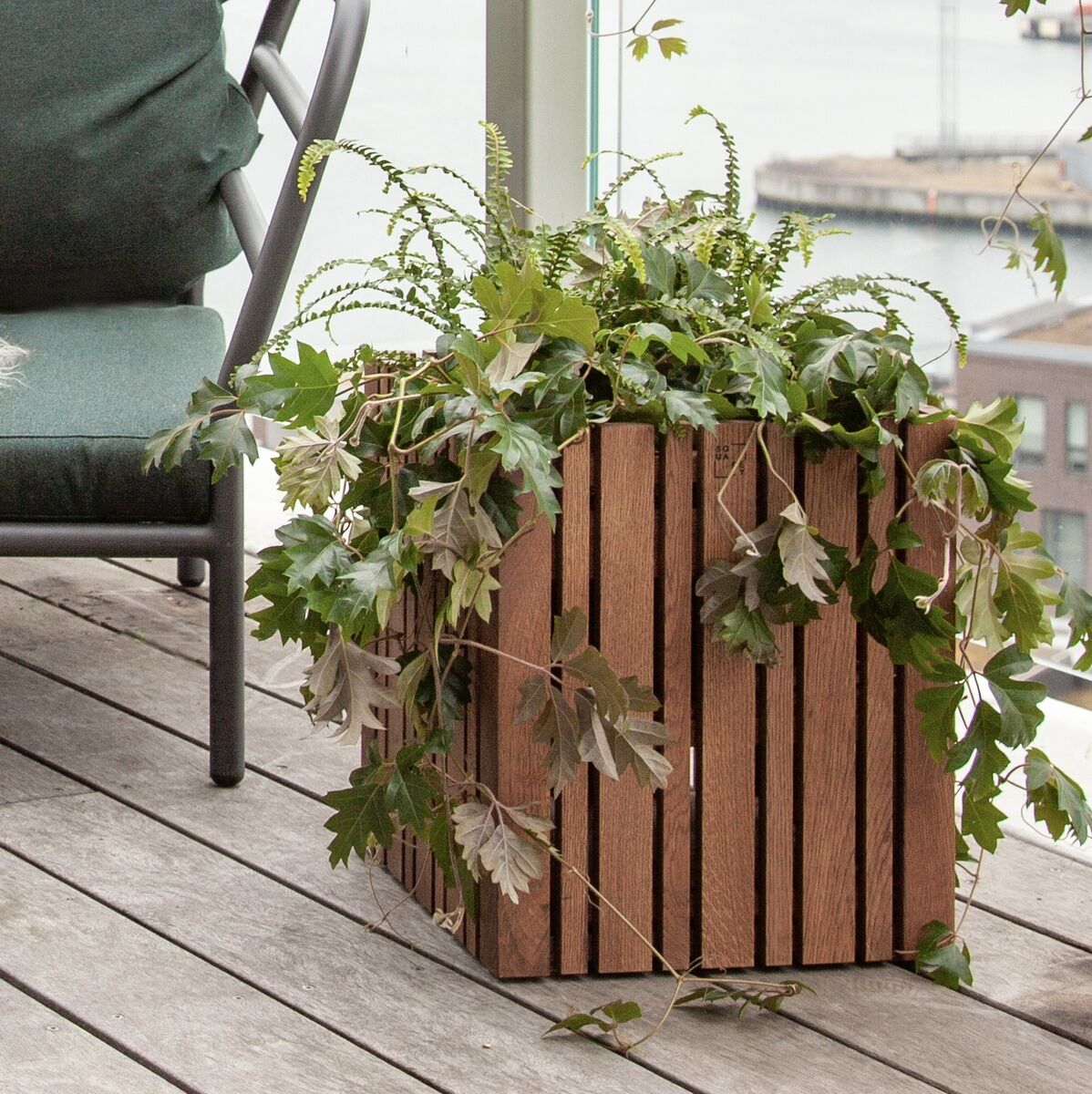 Ein großes rechteckiges Pflanzgefäß »GrowMORE« aus vertikalen Holzlamellen steht auf einer holzverkleideten Terrasse. Es ist mit verschiedenen grünen Pflanzen und Weinblättern bepflanzt. Im Hintergrund befinden sich ein grünes Sitzkissen, ein runder Tisch und ein modernes Outdoor-Sofa. Der Blick erstreckt sich weiter über eine Balustrade hinweg auf eine flussnahe städtische Landschaft.