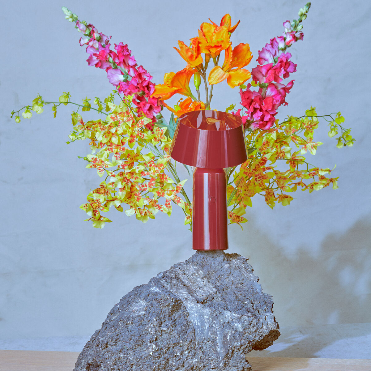 Eine moderne Leseleuchte »Bicoca« in Rot steht vor einem Hintergrund aus farbenfrohen Blumen und grauem Stein. Die Lampe hat ein minimalistisches und elegantes Design und wird von verschiedenen bunten Blumenarrangements umgeben.