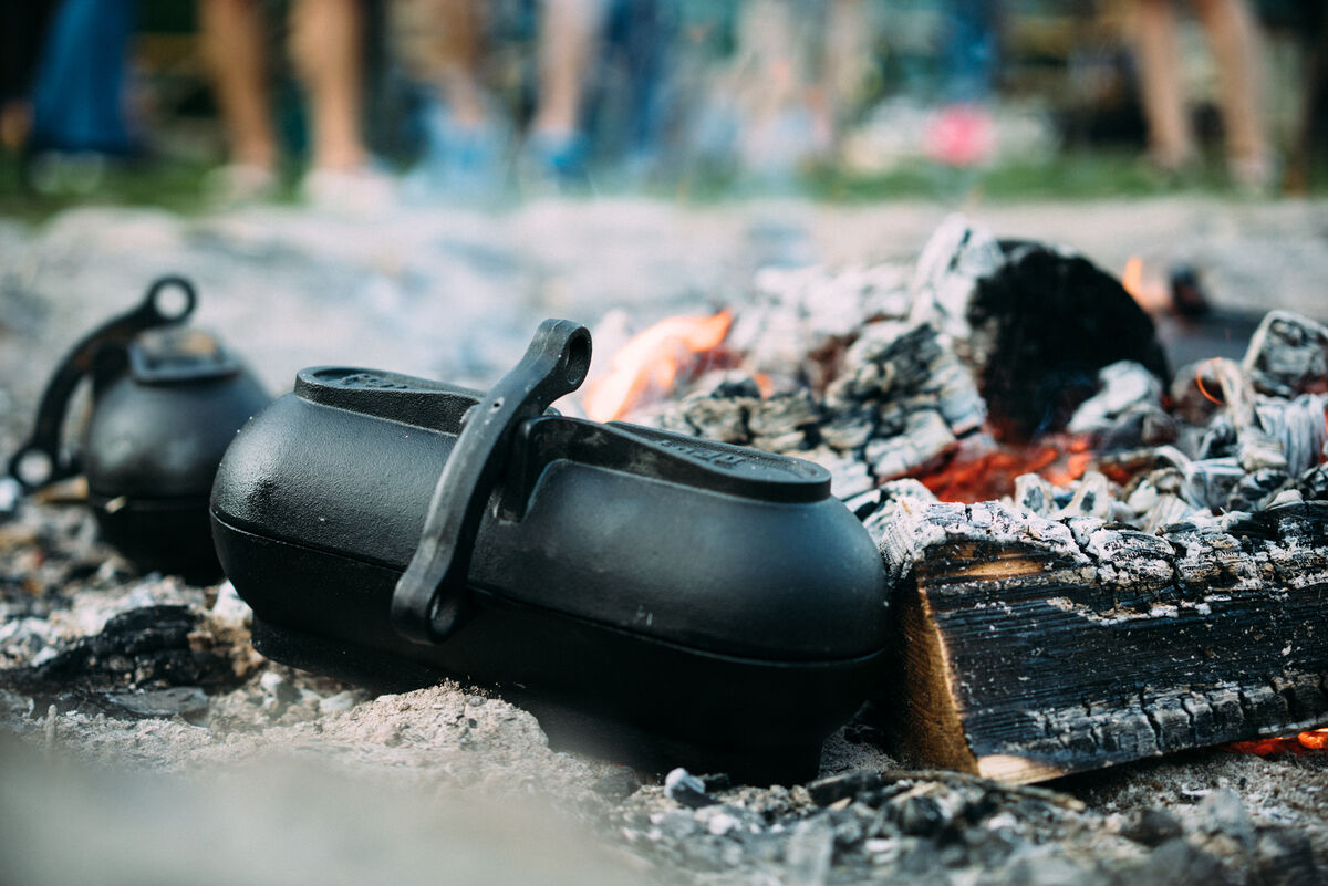 Schwarzer Kartoffelbräter aus Gusseisen über einem rauchenden Holzfeuer mit glühenden Kohlen, umgeben von rustikaler Natur.