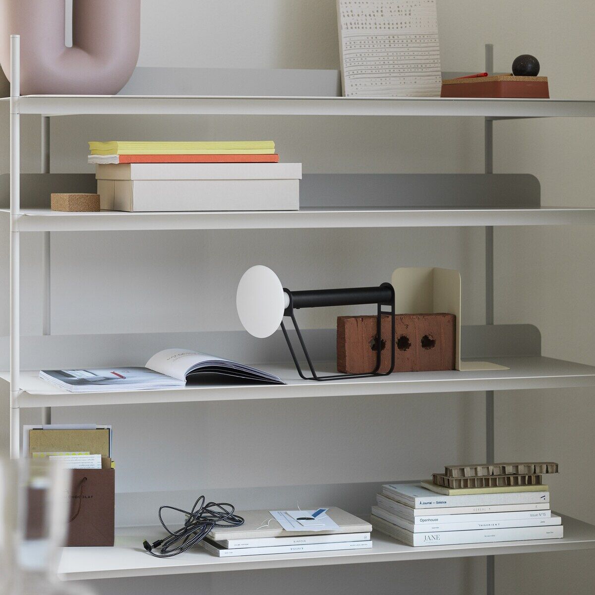 Ein Bücherregal mit mehreren Artikeln, darunter eine tragbare schwarze und weiße Lampe namens »Piton«, Bücher, Pflanzen und Deko-Objekte. Die Lampe steht im mittleren Regal und hat ein modernes, minimalistisches Design.