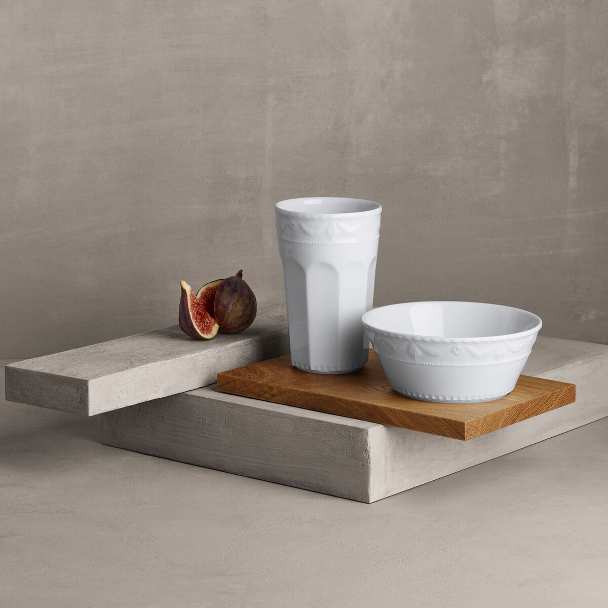 Abgebildet ist ein Müsli-Gourmet-Set »Kurland« von KPM, bestehend aus einer weißen Porzellan-Schale und einem weißen Becher, beide auf einem Holzboden präsentiert. Eine halbierte Feige liegt dekorativ daneben. Der Hintergrund ist in neutralen Grautönen gehalten.