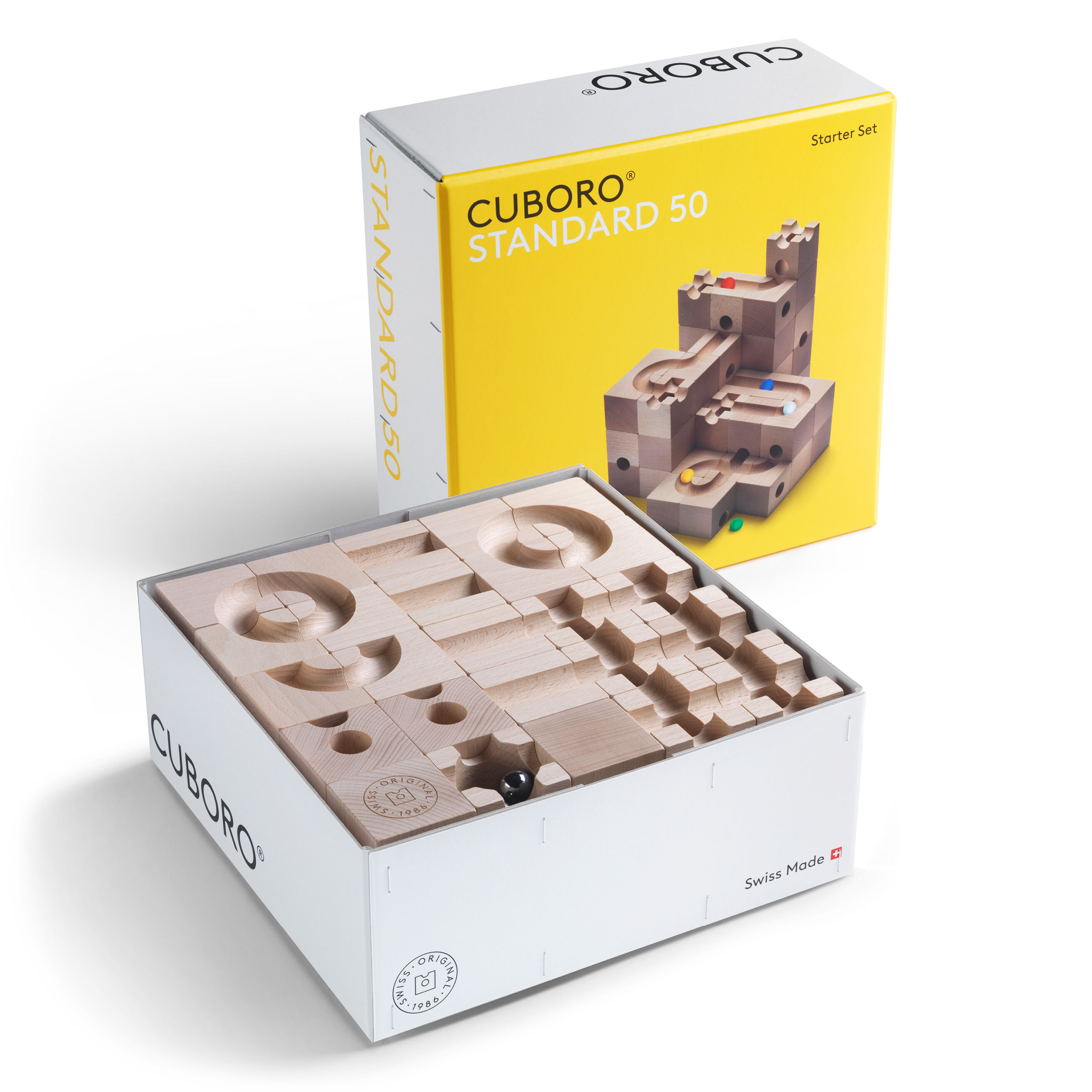 Das Bild zeigt das Cuboro Kugelbahn Starter-Set »STANDARD« mit 50 Elementen. Die geöffnete, weiße Verpackung offenbart präzise gefertigte Buchenholz-Bausteine. Im Hintergrund ist der gelbe, geschlossene Umkarton mit Produktabbildung und Aufschrift zu sehen.
