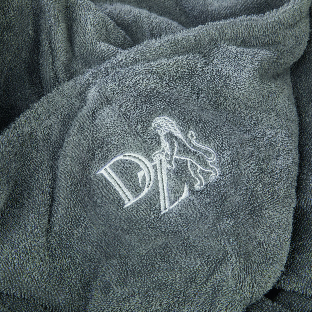 Ein grauer Frottee-Bademantel mit einem gestickten weißen Logo, das einen stilisierten Buchstaben 'DL' und einen Löwen darstellt. Der Stoff des Bademantels wirkt weich und bequem.