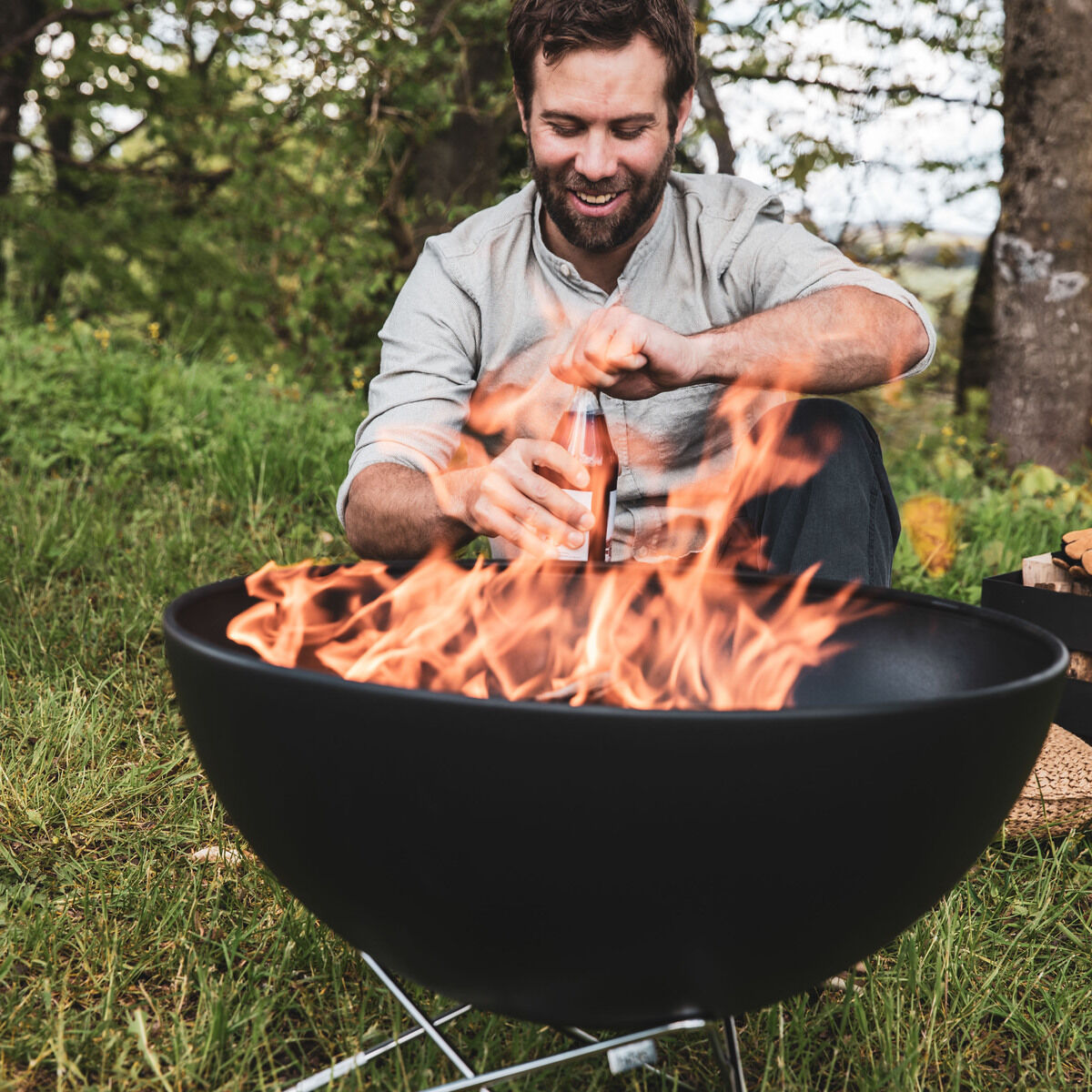 Ein Mann sitzt im Freien vor einer brennenden Feuerschale »BOWL 57« und hält eine Bierflasche. Die Feuerschale steht auf grasbewachsenem Boden mit Bäumen im Hintergrund.