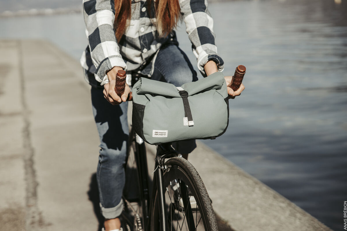 Eine Person in einem karierten Hemd fährt ein Fahrrad entlang eines Gewässers mit einer grauen Lenkertasche »Piha«. Die Tasche ist an den Fahrradlenker montiert und mit einem schwarzen Riemen gesichert.