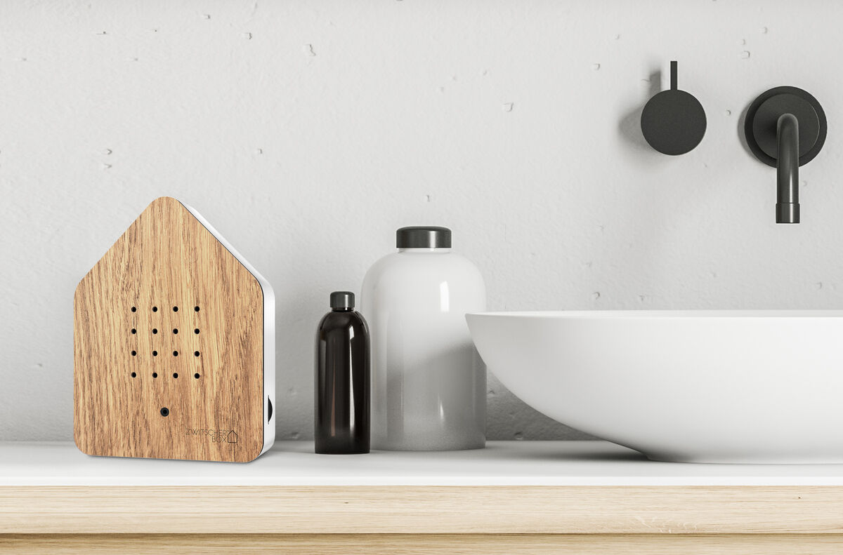 Die Zwitscherbox steht auf einer hellen Holzoberfläche im Badezimmer, neben einer weißen Schale und Flüssigkeitsbehältern. Im Hintergrund sind graue Wände und schwarze Armaturen erkennbar.