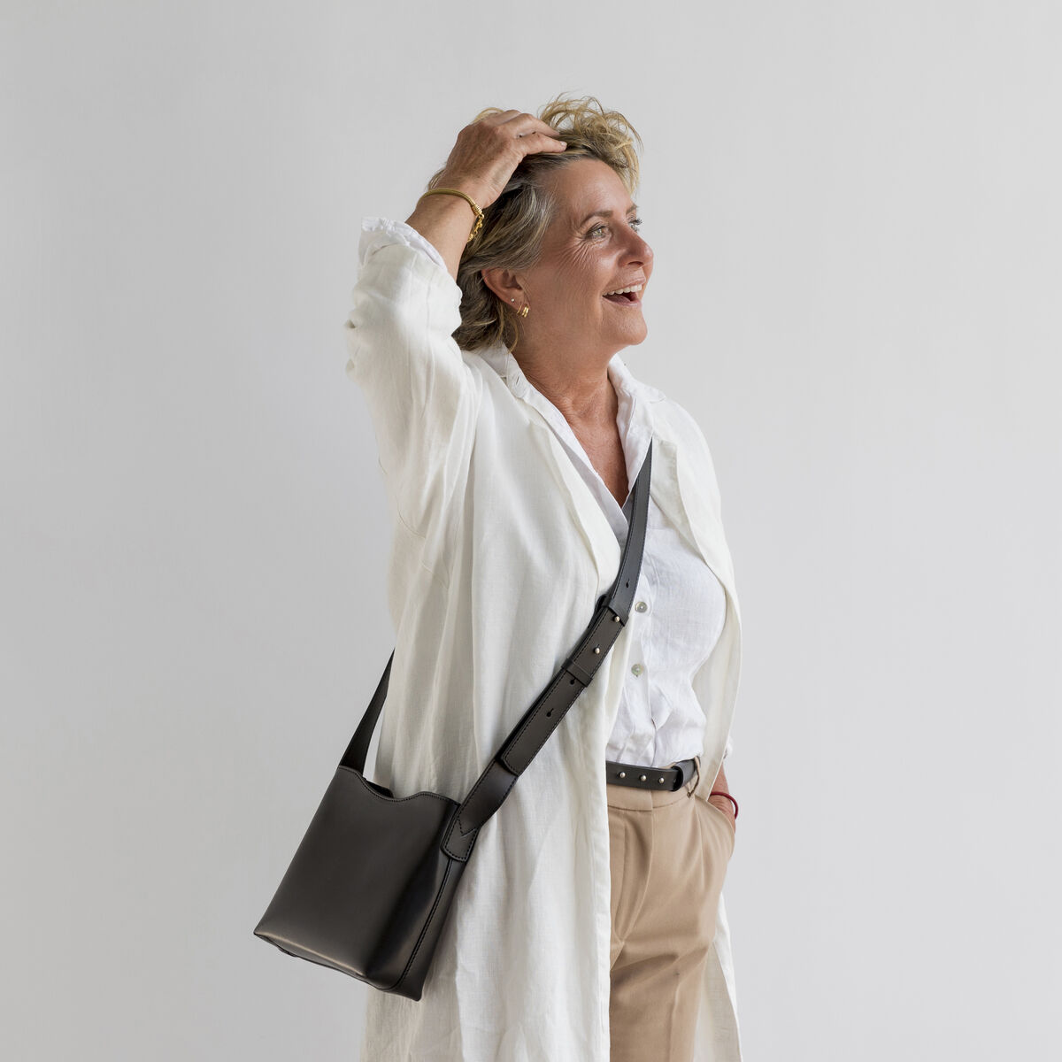Eine Frau trägt eine schwarze Schultertasche aus Kernleder. Sie ist in einem eleganten, weißen Outfit, bestehend aus einem weißen, langärmeligen Oberteil und beige Hose, gekleidet. Der Hintergrund ist schlicht und weiß.