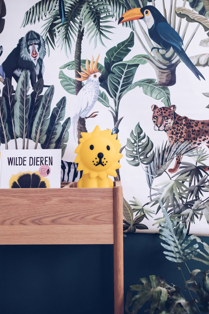 Ein Nachtlicht in Form eines Löwen, das auf einem hölzernen Regal in einem Kinderzimmer steht. Im Hintergrund ist eine Tapete mit verschiedenen Dschungeltieren und Pflanzen zu sehen, darunter ein Leopard, ein Affe und verschiedene Vögel. Auf dem Regal liegt ein Kinderbuch mit dem Titel 'Wilde Dieren'.