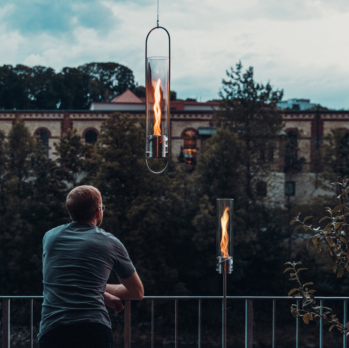 Ein Mann lehnt sich auf einem Balkon, betrachtet die beleuchteten SPIN Hängesysteme, die wie Feuerflammen in modernen Glaszylindern erscheinen. Der Hintergrund zeigt eine alte architektonische Struktur und bewölkten Himmel.