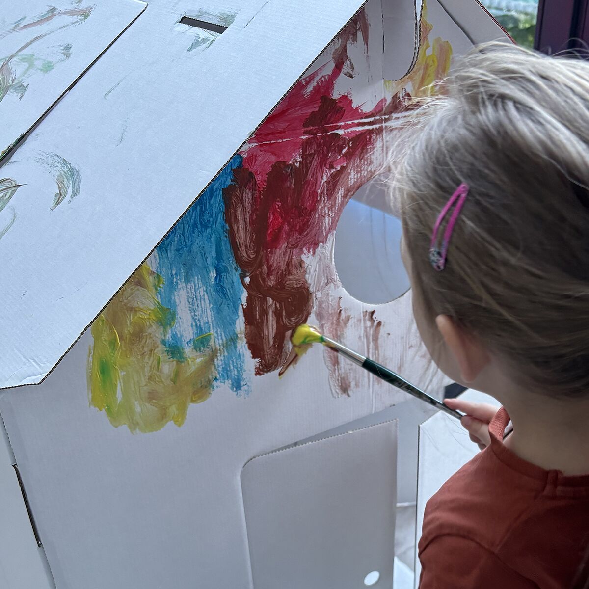 Ein Kind bemalt mit einem Pinsel ein großes, weißes Papphaus der Marke bibabox. Das Haus aus stabiler Wellpappe zeigt bunte Farbflecken in Gelb, Rot, Blau und Grün. Im Vordergrund ist der Rücken des Kindes mit Haarspange und rotem Shirt zu sehen. Im Hintergrund fällt Tageslicht durch ein Fenster.
