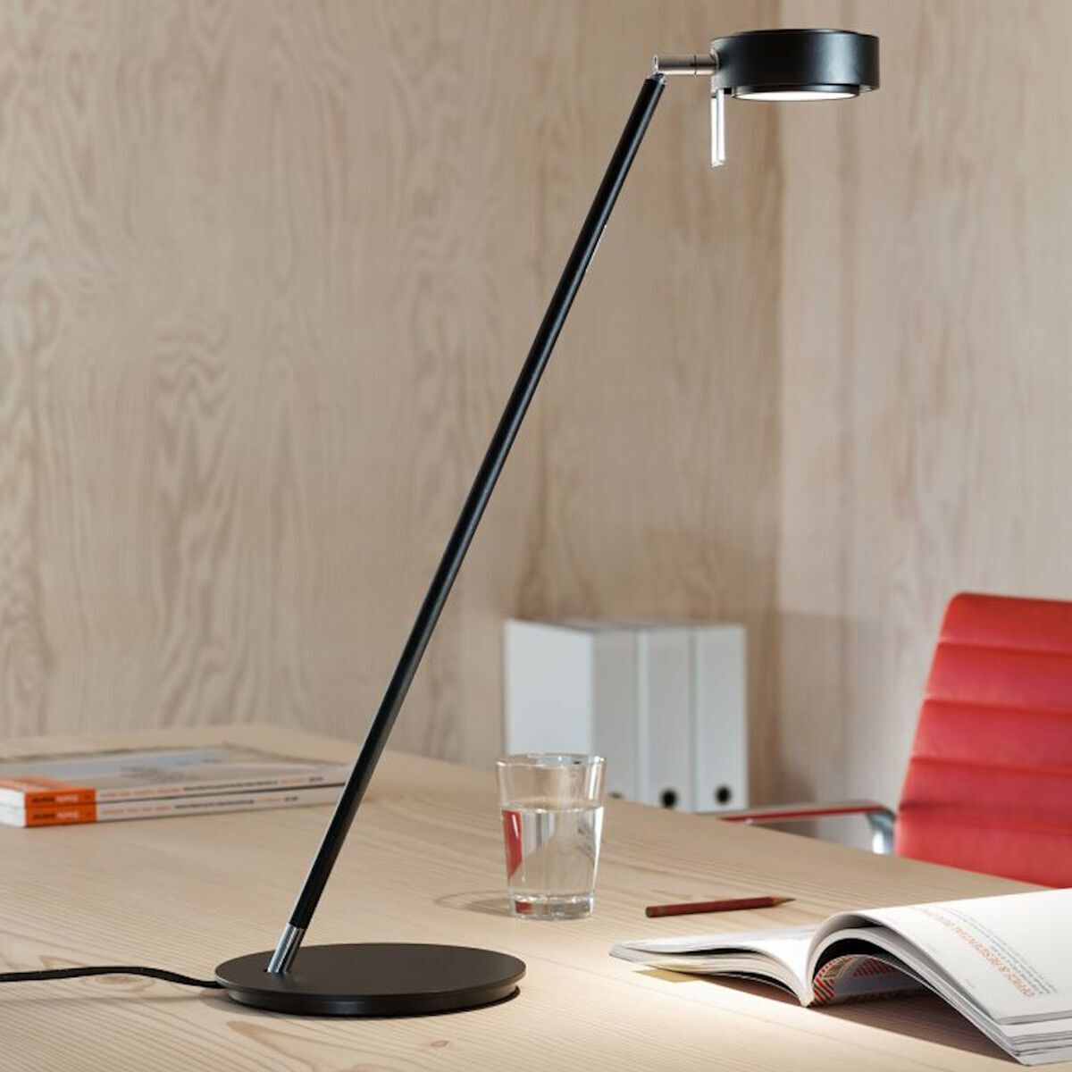 Eine moderne, schwarze Tischleuchte namens 'Pure' steht auf einem hellen Holztisch. Daneben befindet sich ein Glas Wasser, einige Papierstapel und ein roter Stuhl. Im Hintergrund sind eine Wand in Holzoptik und ein weißer Aktenordner zu sehen.