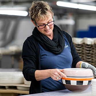 Frau mit Brille und kurzem Haar inspiziert eine weiße Schüssel mit orange-schwarzem Muster in einer Werkstatt