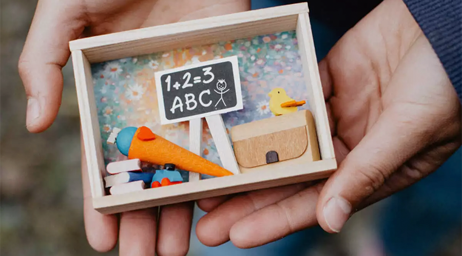 Hände halten eine kleine Holzkiste mit Miniatur-Schultüte, Schulranzen, Tafel mit Rechnung '1+2=3' und ABC, sowie kleinen Büchern als Einschulungsgeschenk