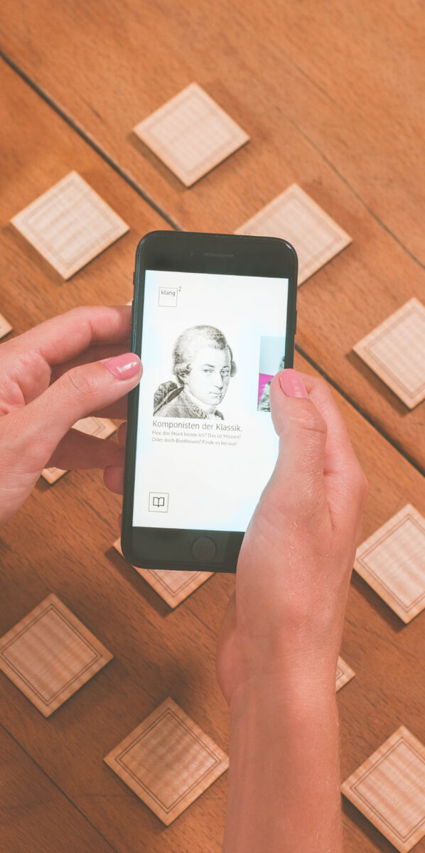 Ein Smartphone mit einer App, die Geigenbauer darstellt, wird von zwei Händen gehalten. Im Hintergrund sind mehrere kleine Holzplättchen auf einem Holztisch angeordnet.