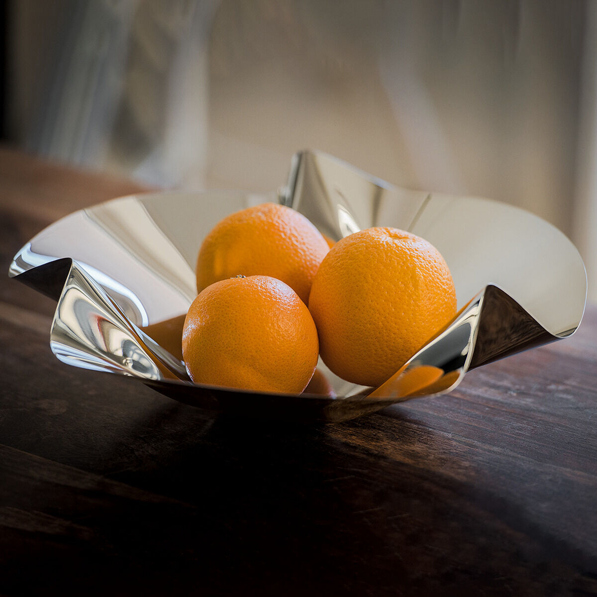 Schale »Margarethe« aus Edelstahl mit organischer Form, gefüllt mit drei Orangen, auf einem dunklen Holztisch platziert. Die Schale reflektiert das Licht und erzeugt eine glänzende und moderne Ästhetik.