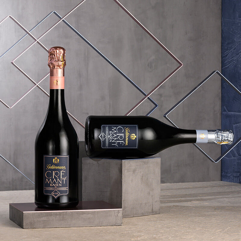 Ein Crémant Spezialitäten Paket inklusive Neoprenmanschetten. Zwei Flaschen Crémant, eine mit einer rosafarbenen Kapsel und die andere mit einer silberfarbenen Kapsel, steht auf einem grauen Hintergrund mit geometrischem Muster.