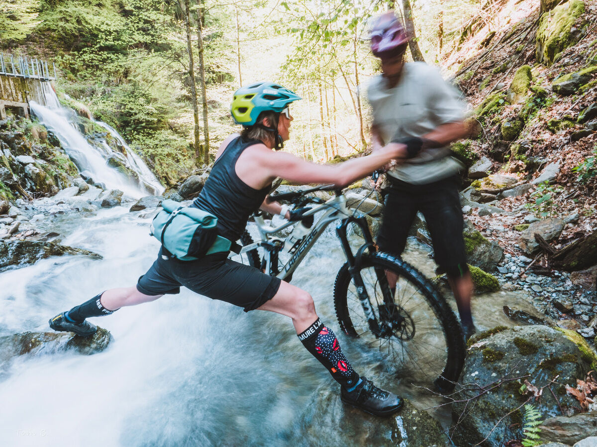 Zwei Personen überqueren auf Mountainbikes einen Fluss, umgeben von Wald und einem Wasserfall im Hintergrund. Die Person vorne trägt eine Lenkertasche vom Modell 'Piha' und zeigt sportliche Kleidung und Ausrüstung.