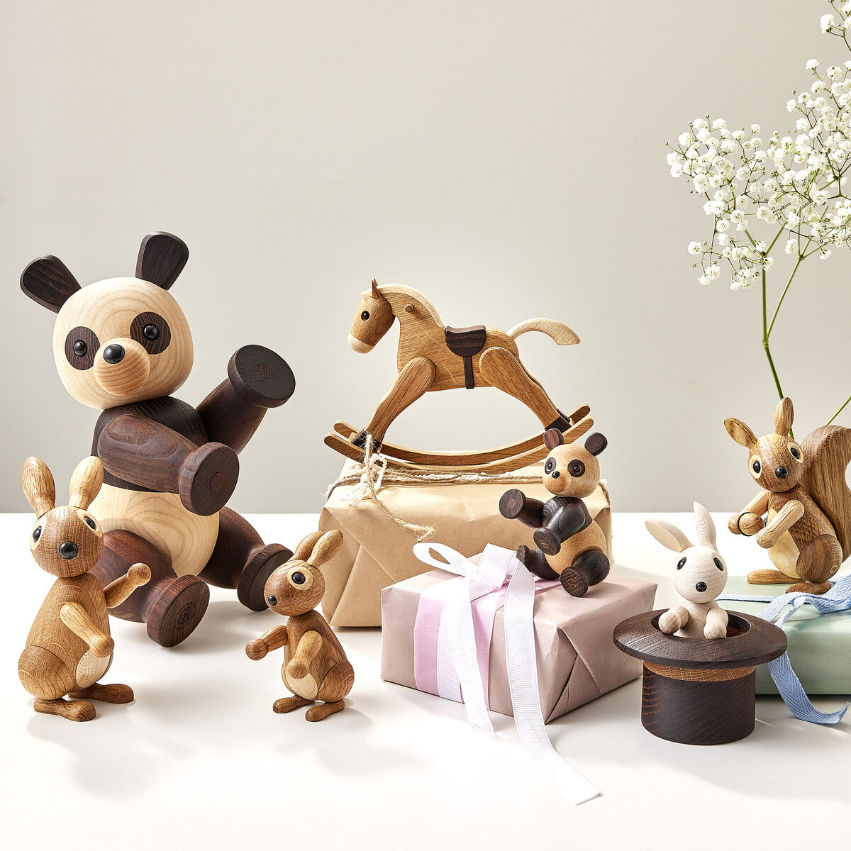 Mehrere handgefertigte Holzfiguren, darunter ein Schaukelpferd, verschiedene Tiere wie Panda, Hasen und Eichhörnchen sowie liebevoll verpackte Geschenke mit Schleifen, sind auf einem Tisch neben zarten weißen Blumen arrangiert.