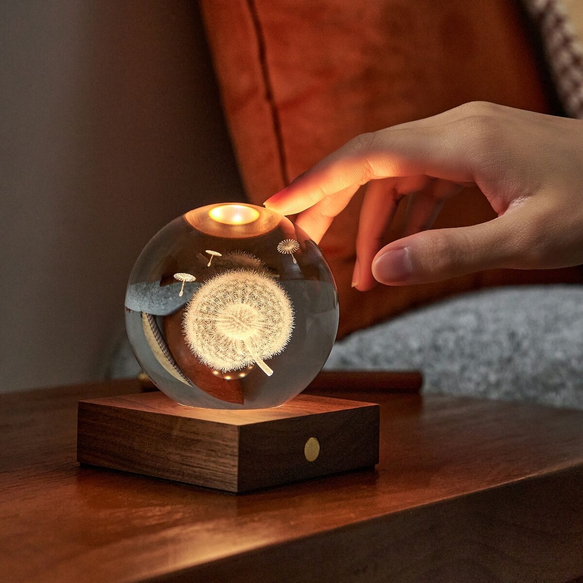 Auf einem Holztisch steht die Lampe »Amber Light« von Gingko Design Ltd mit rundem Kristallglas und filigranem Dandelion-3D-Lasermotiv. Ein warmes Licht scheint aus dem edlen Sockel aus Walnussholz. Eine Hand aktiviert gerade per Touch den Kupferknopf, Couch und Kissen im Hintergrund.