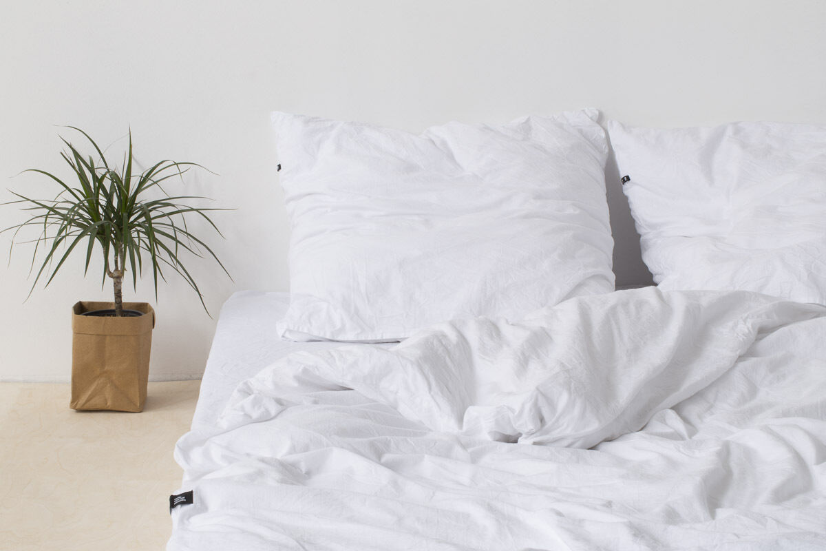 Eine minimalistische Bettwäsche namens „Pure“ in Weiß, arrangiert auf einem Bett mit weißen Kissen und Bezügen. Daneben steht eine grüne Zimmerpflanze in einem braunen Papierübertopf auf dem Boden.