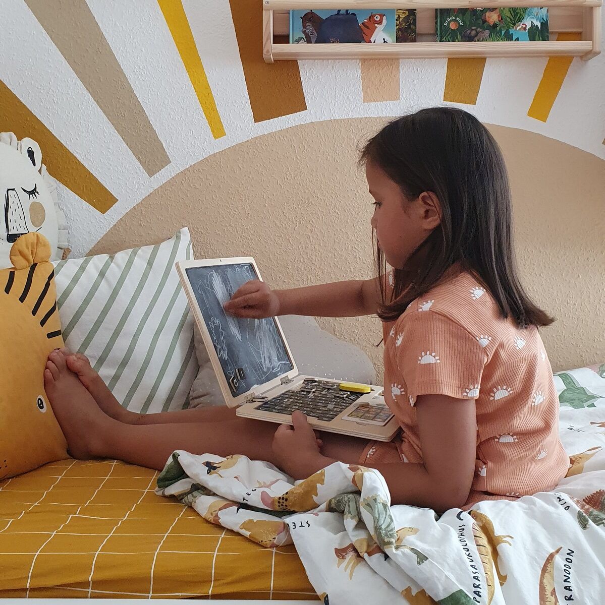 Ein Mädchen sitzt auf einem Bett in einem Kinderzimmer und spielt mit einem Holz Laptop mit Magnettafel. Im Hintergrund sind Bücher, ein Stofftier und eine gelb-weiße Wand mit einem gemalten Regenbogen zu sehen.