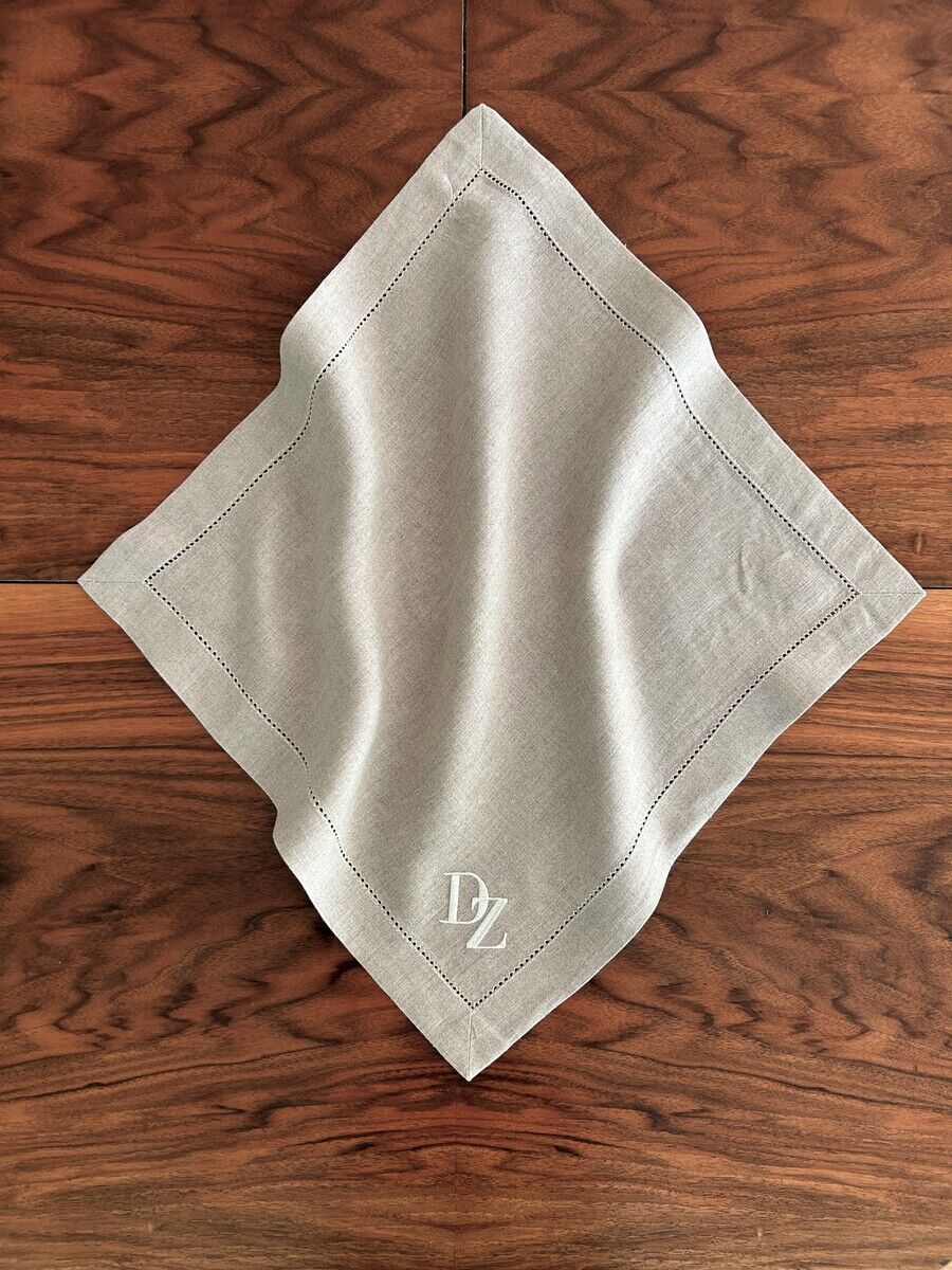 Leinen-Serviette in Naturfarbe mit Monogramm. Die Serviette ist kunstvoll auf einem Holztisch drapiert und zeigt eine dezente, elegante Stickerei.