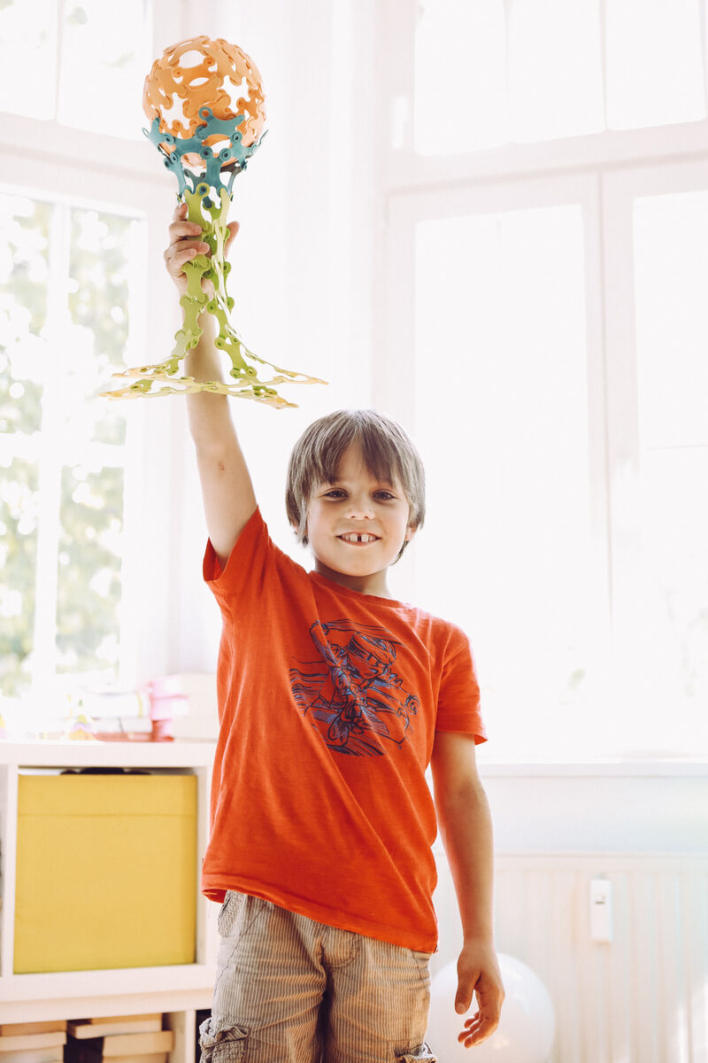 Ein Kind mit einem orangen T-Shirt hält ein farbenfrohes selbstgebautes Modell aus dem Spiel 'Binabo' in die Höhe. Das Bild zeigt Freude und Kreativität in einem hellen, freundlichen Raum.