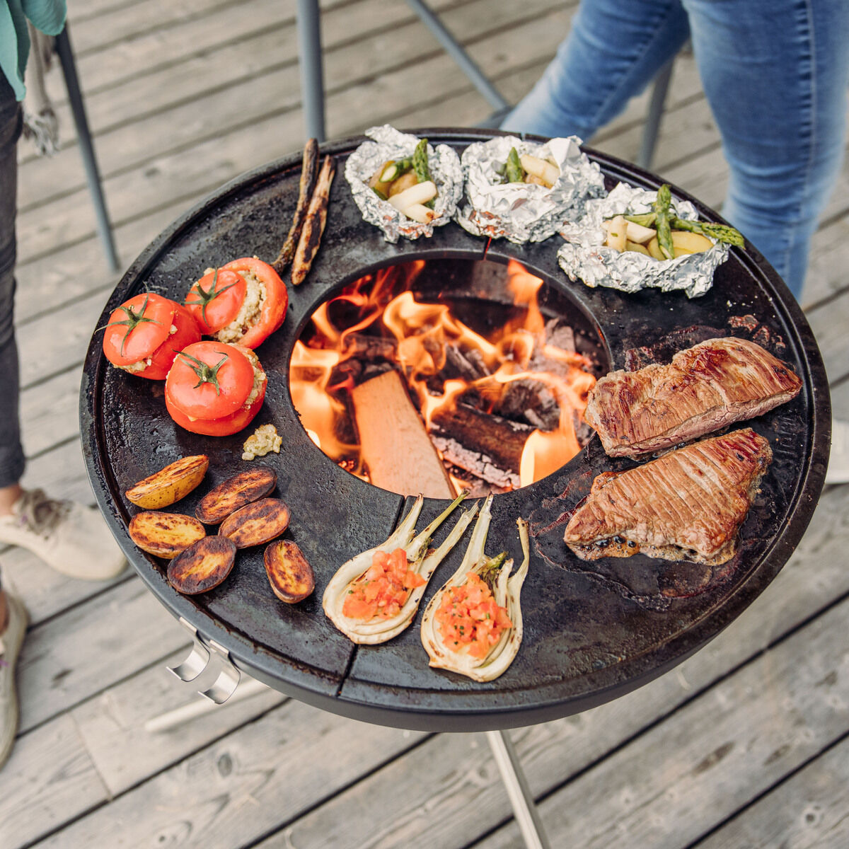 Eine Gruppe von Personen steht um den »BOWL 57« Plancha-Grillset mit einer kreisförmigen Grillplatte. Auf dem Grill liegen Tomaten, Fleischstücke, Kartoffeln und Gemüse in Folie. Im Zentrum brennt ein Holzfeuer. Der Grill steht auf einer hölzernen Terrasse.