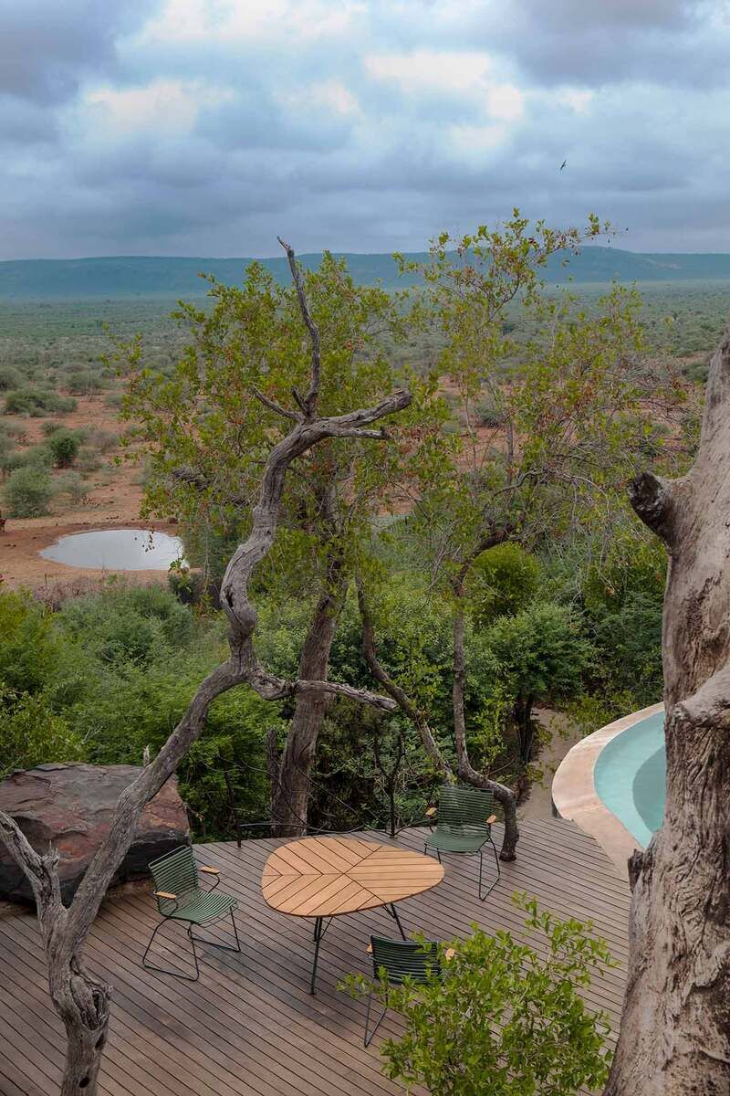 Ein runder Gartentisch aus Holz, umgeben von grünen Stühlen, steht auf einer Holzterrasse. Im Hintergrund befinden sich Bäume, eine Naturfläche und ein Pool. Der Himmel ist bewölkt.