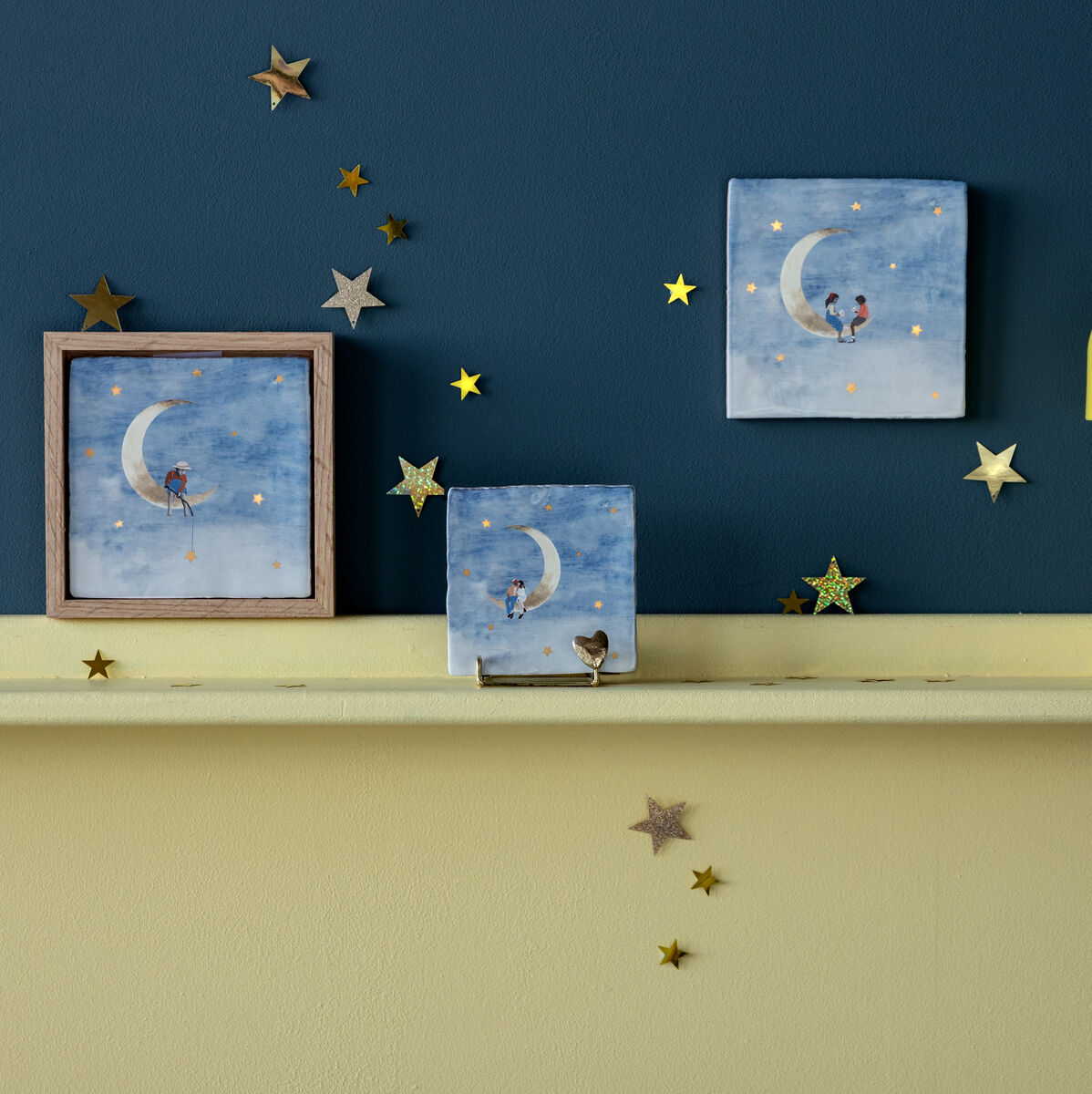 Ein Regal mit einer gelben Lampe und drei Keramikfliesen, die eine Szene von einem Mond mit Sternen und Figuren zeigen. Die Wand dahinter ist blau und das Regal ist gelb. Kleine goldene Sterne sind als Dekoration verteilt.