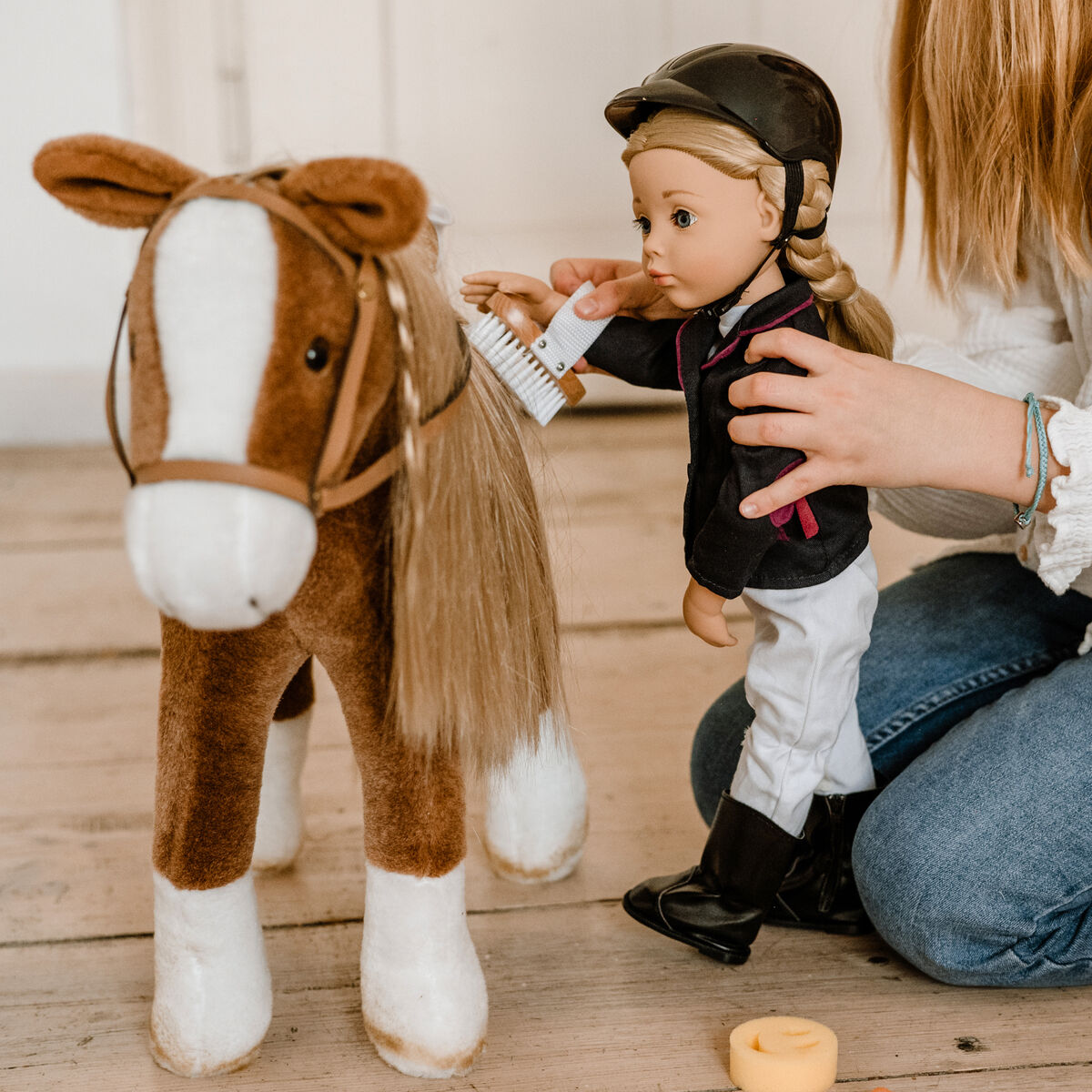 Ein Kind spielt drinnen auf hellem Holzboden mit einem braunen Plüschpferd namens Kämmpferd Brown Beauty und einer Puppe im Reitoutfit. Das Kind bürstet gemeinsam mit der Puppe die Mähne des Pferdes. Im Vordergrund sind Zubehörteile zu sehen.