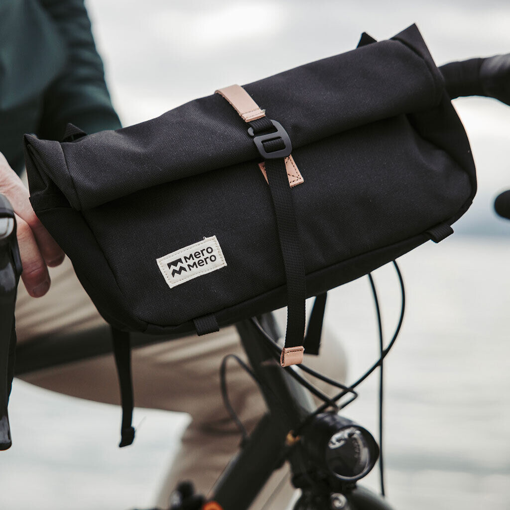 Schwarze Fahrradtasche »Piha« aus wasserdichtem Material mit Lederdetails und Schnallenbefestigung, montiert an einem Fahrradgepäckträger. Eine Person im Hintergrund hält den Lenker.