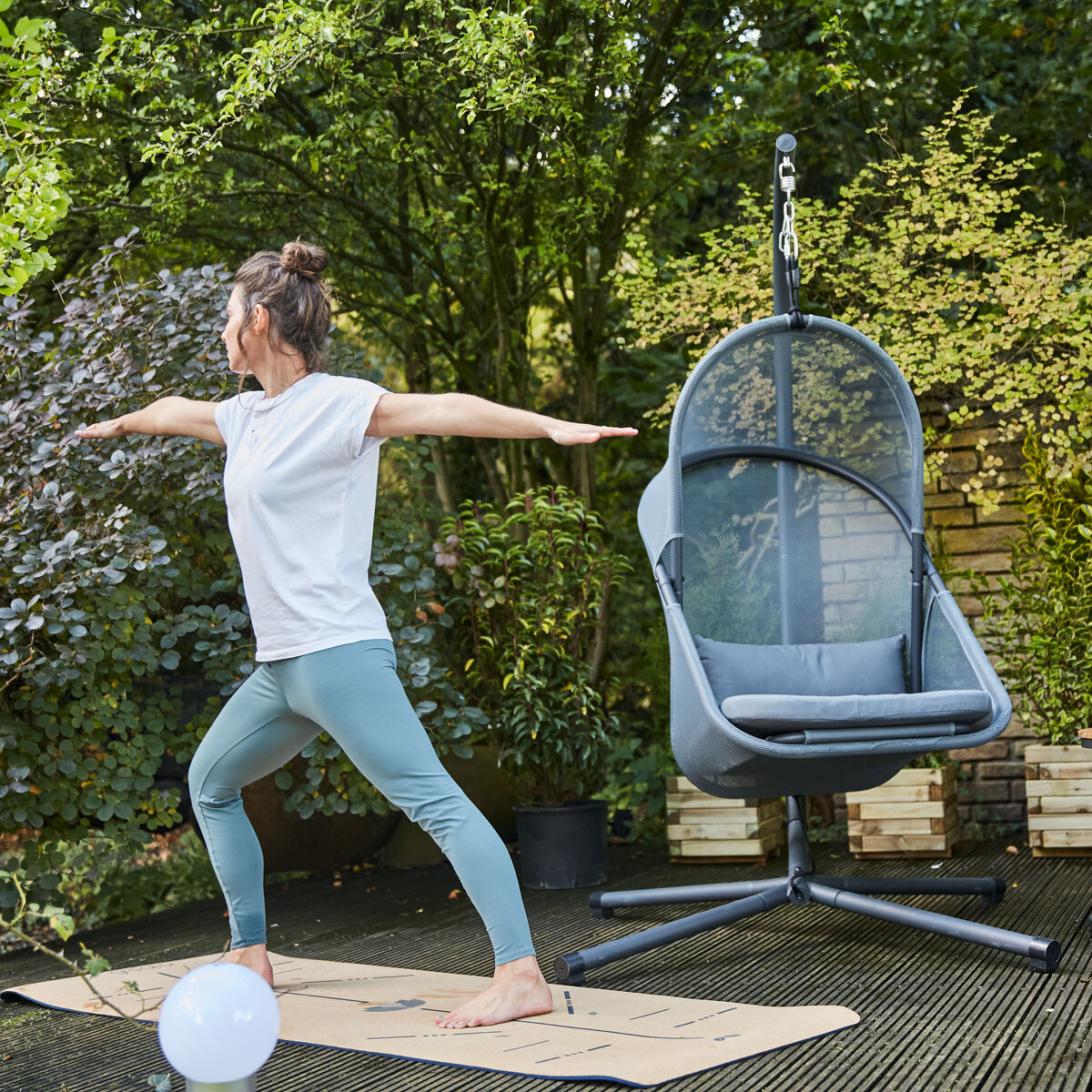 Eine Frau in Yoga-Position auf einer Terrasse neben einem modernen grauen Hängesessel »Yobaya«, umgeben von grünen Pflanzen und Gartenmöbeln.