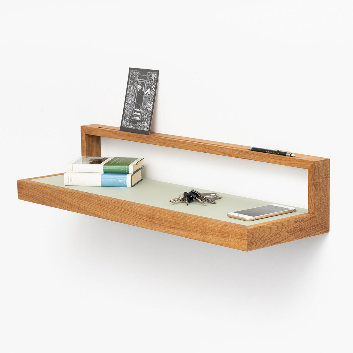 Schwebendes Ablagebord „buck“ in Eiche und Pistaziengrün, rechteckig mit klaren Linien. Auf dem Regal liegen Schlüssel, Buch, Notizbuch, Stift und Smartphone. Hinten ist eine eingelassene Leiste. Das Wandregal wirkt modern und minimalistisch an einer weißen Wand.