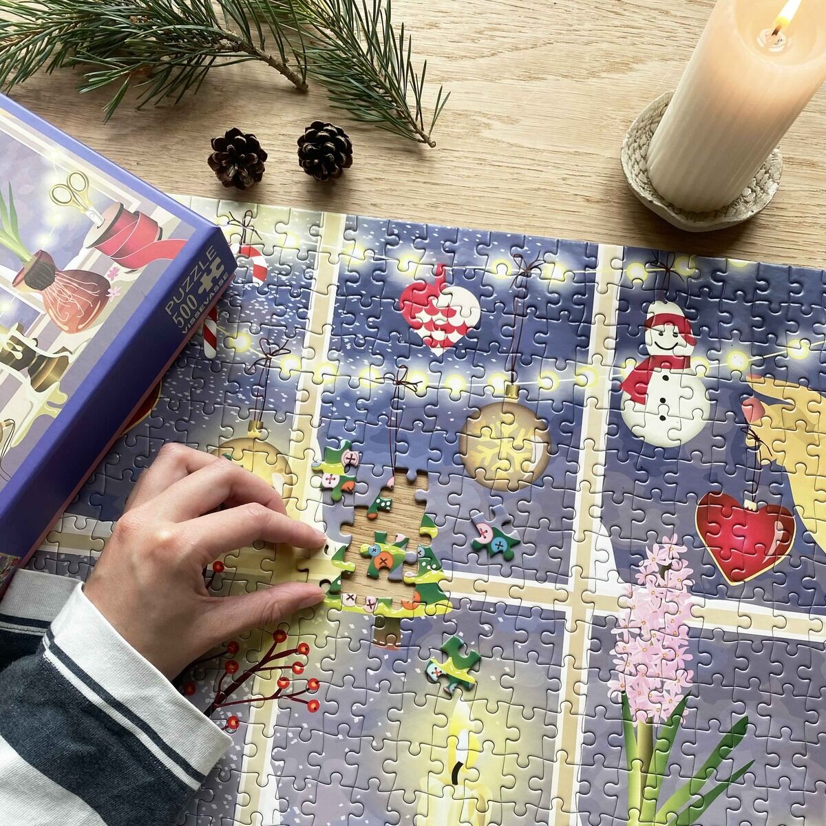 Ein 500 Teile Puzzle »Weihnachtsfenster« von VISSEVASSE liegt teils fertig auf einem Holztisch. Zu sehen sind dekorative Motive wie Schneemann, Weihnachtsbaum und Fensterdekoration. Eine Hand setzt gerade Puzzleteile ein, daneben eine brennende Kerze, Kieferzweige und Tannenzapfen.