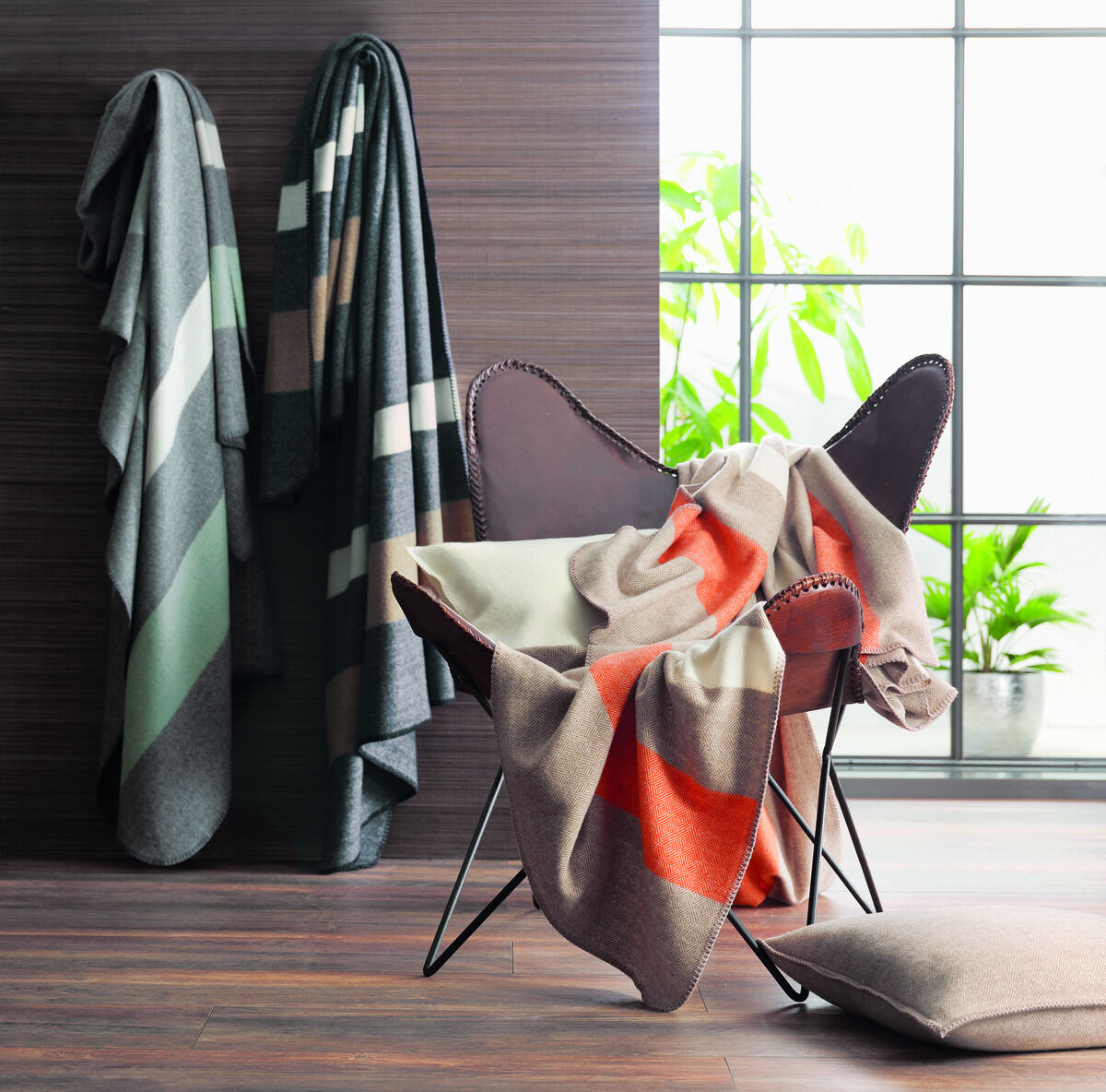 Eine moderne und stilvolle Plaid »Liverpool«-Decke hängt über einem anthrazitfarbenen Stuhl, platziert vor einem Fenster mit grünen Pflanzen. Die Decke hat geometrische Muster in Grautönen, Weiß, Orange und Grün. Eine weitere, ähnliche Decke ist im Hintergrund zu sehen, aufgehängt an einem dunklen Holzwandpaneel.