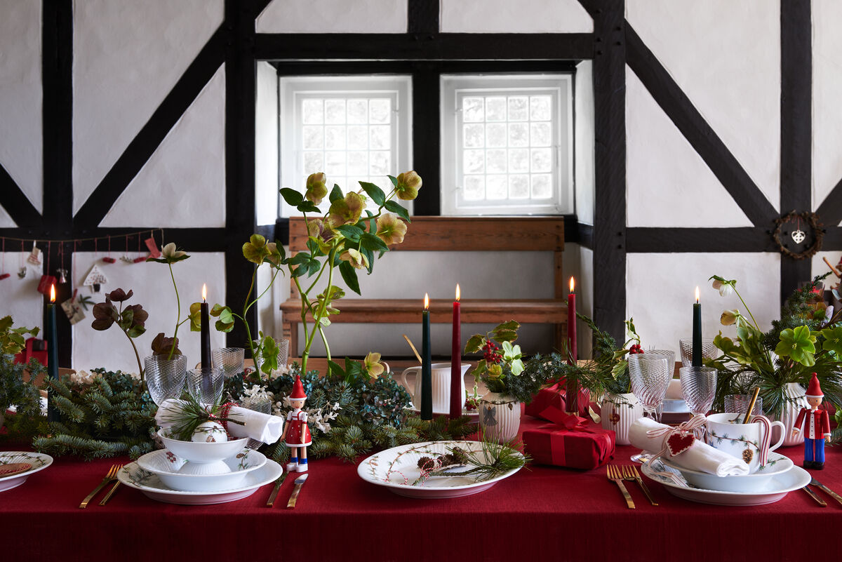 Eine festliche Weihnachtstafel in einem traditionellen Raum. Die Tischdecke ist rot und mit geschmückten Tannenzweigen, brennenden Kerzen und elegantem weißem Geschirr mit floralen Mustern dekoriert. Auf der linken Seite steht ein Nussknacker und servierte Platten. Im Hintergrund sind Sprossenfenster und eine rustikale Bank zu sehen.