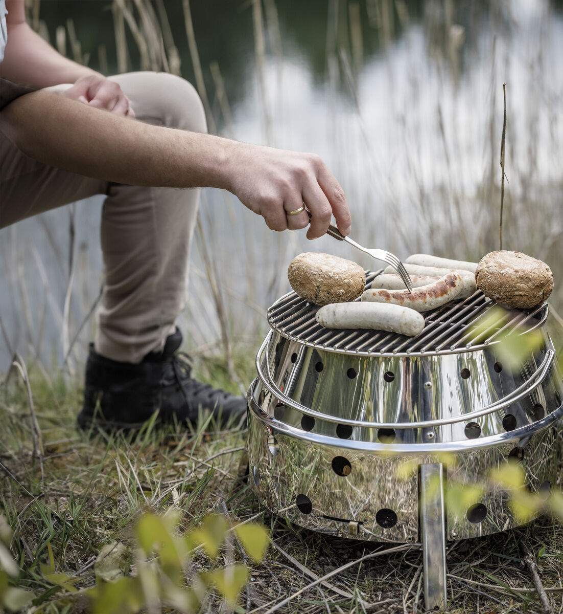Eine Person grillt Würstchen und Brot auf einem runden Edelstahlgrill des Modells 'Atago', der im Freien neben einem See aufgestellt ist. Gras und Blätter sind im Vordergrund zu sehen.