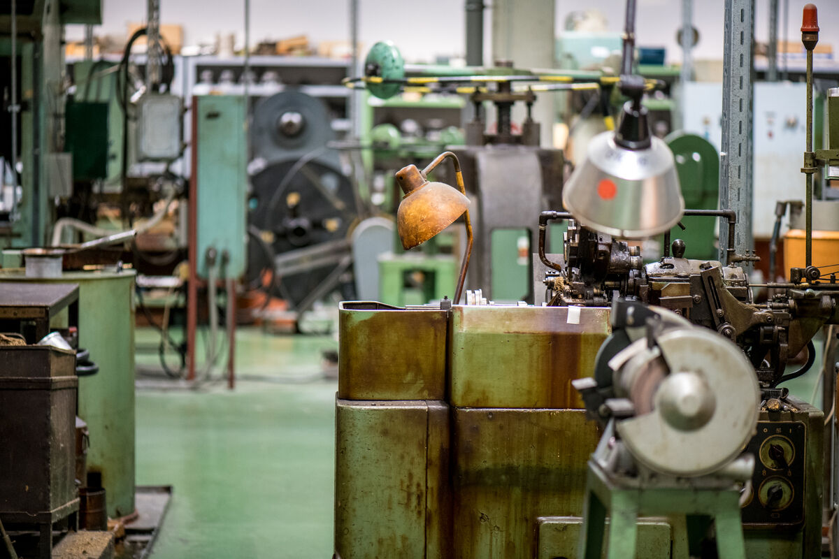 Eine industrielle Werkstatt mit verschiedenen Maschinen und Werkzeugen, darunter dicke grüne Arbeitsstationen und eine alte Lampe, die dem Raum ein Retro-Ambiente verleiht. Verschiedene Metallstrukturen und grüne Farbgebung dominieren das Bild.