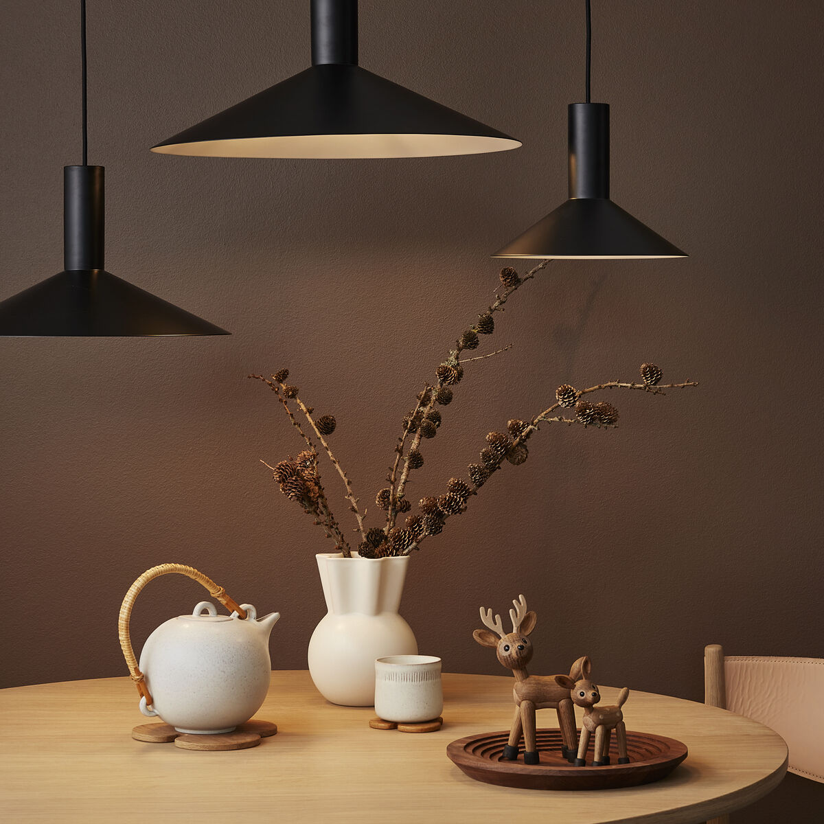 Ein runder Holztisch steht vor einer braunen Wand und ist mit minimalistischer Dekoration gestaltet: Eine cremefarbene Teekanne mit Becher, eine weiße Vase mit trockenen Zweigen und eine Holzfigur in Form eines Rentiers auf einem runden Holztablett. Drei schwarze Pendelleuchten hängen von der Decke und erzeugen ein gemütliches Lichtambiente.