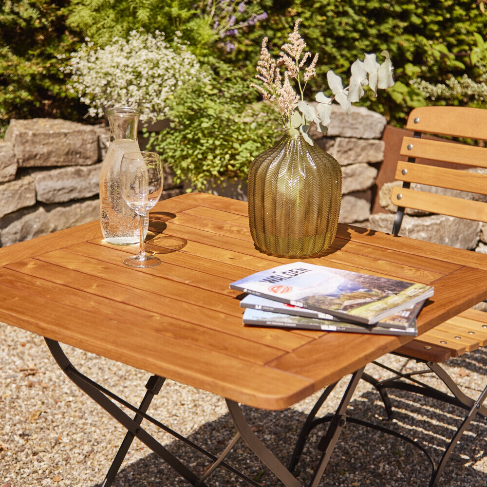 Ein quadratischer Klapptisch aus Holz mit Metallbeinen steht in einem Gartenbereich mit grünen Pflanzen im Hintergrund. Auf dem Tisch stehen eine Vase mit Blumen, ein Glas Wasser und einige Zeitschriften. Der Tisch ist auf einem Kieselboden platziert und von einer Holzmauer umgeben.