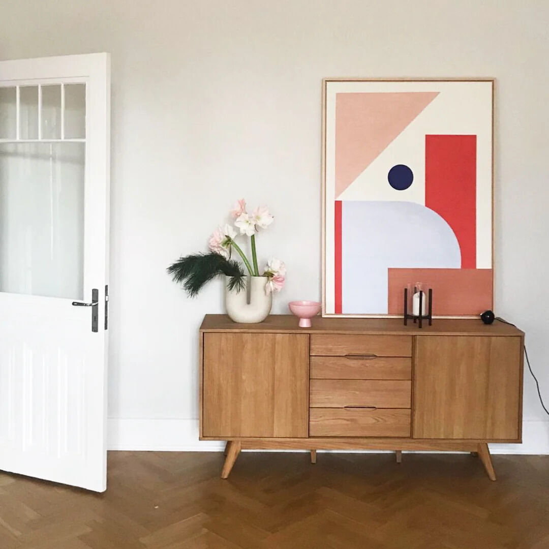 Ein modernes Leinwandbild mit abstrakten, geometrischen Formen in Pastellfarben hängt über einer Holzkommode. Auf der Kommode stehen eine Vase mit Blumen, ein kleines rosa Gefäß und zwei dekorative Bücher. Der Raum wirkt hell und minimalistisch.