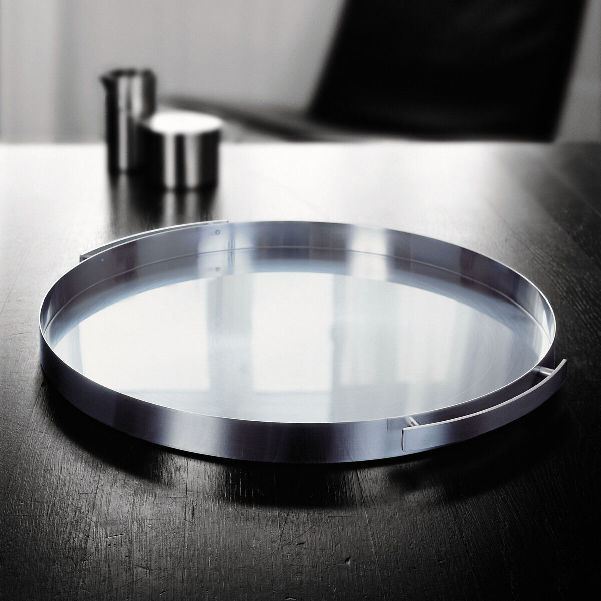 Ein modernes, rundes Serviertablett aus Edelstahl von Arne Jacobsen auf einem dunklen Tisch, mit einem minimalistischen Design. Im Hintergrund sind unscharf ein schwarzer Stuhl und weitere metallische Gegenstände zu erkennen.