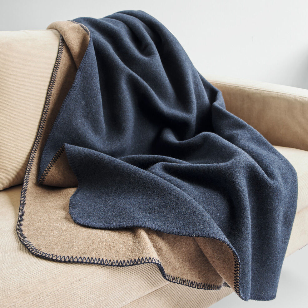 Die Merinowolldecke »DUO« von Eagle Products liegt dekorativ und weich auf einem hellen Sofa. Eine Seite ist dunkelblau, die andere braun, der Kammstich ist gut sichtbar. Die Decke wirkt dick, warm und hochwertig, perfekt für gemütliche Stunden im Wohnzimmer.