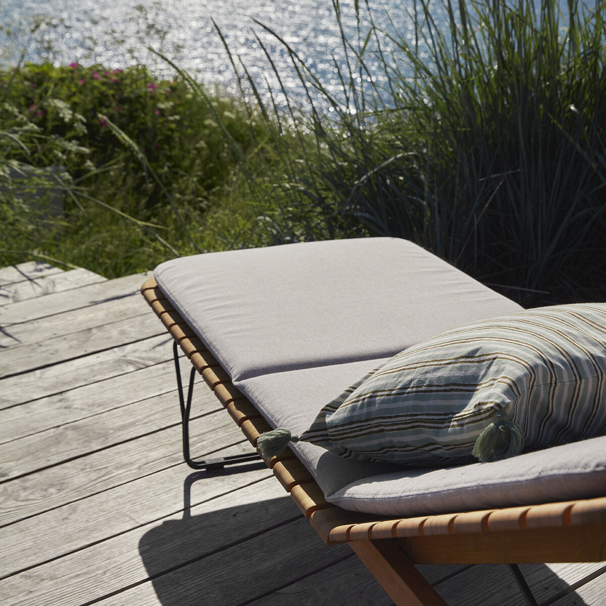 Ein komfortabler Liegestuhl mit weißer Auflage »MOLO« auf einer Holzterrasse mit Blick auf das Meer. Das Sonnenlicht glitzert auf dem Wasser, und im Hintergrund sind Strandhafer und grüne Pflanzen zu sehen. Perfekt für Entspannung und Außenbereich Dekoration.