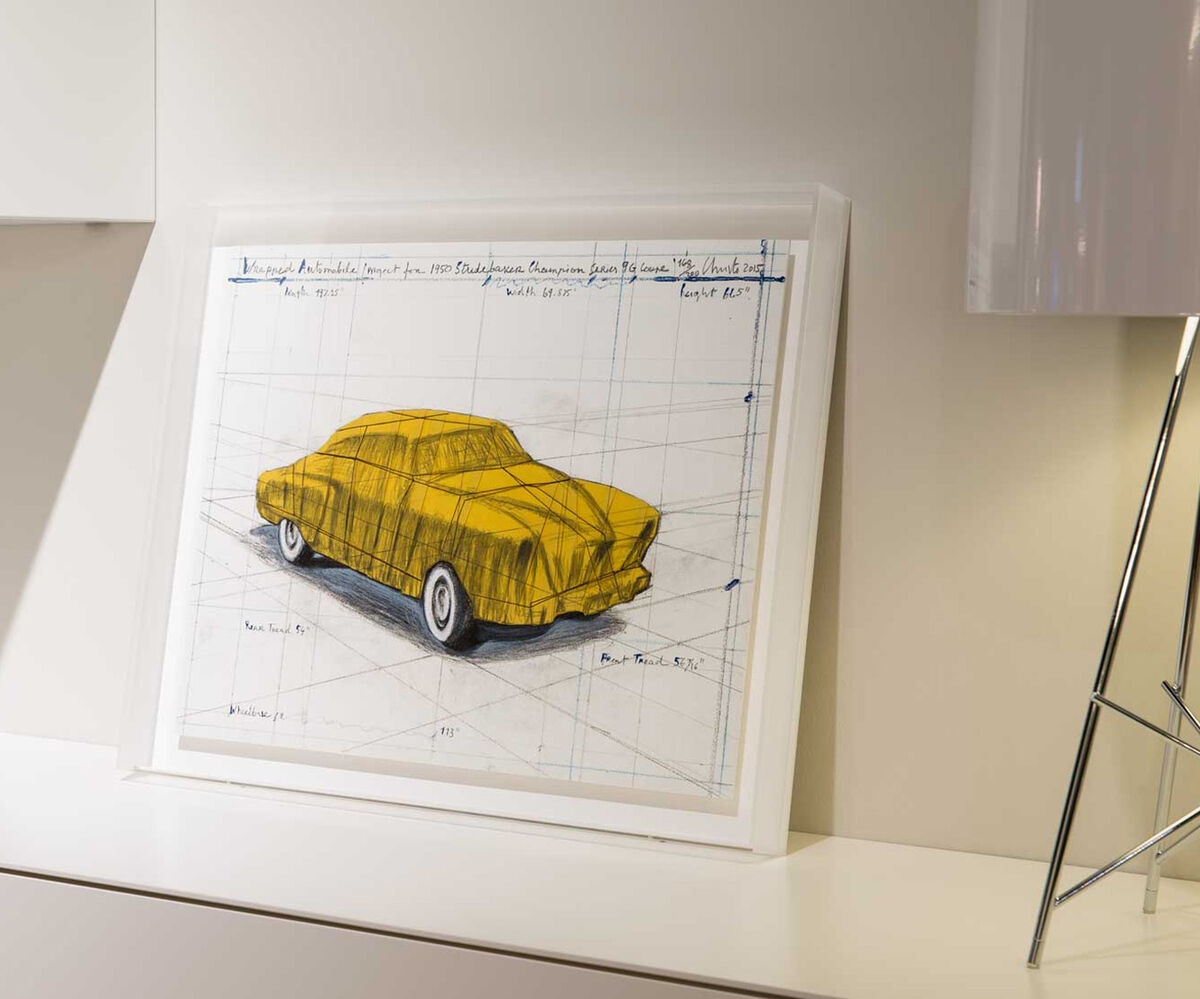 Ein Kunstwerk von Christo zeigt ein eingepacktes Automobil, speziell ein Studebaker, auf Papier dargestellt. Es ist in einer modernen Umgebung, mit einem dominierenden Gelbton, präsentiert.